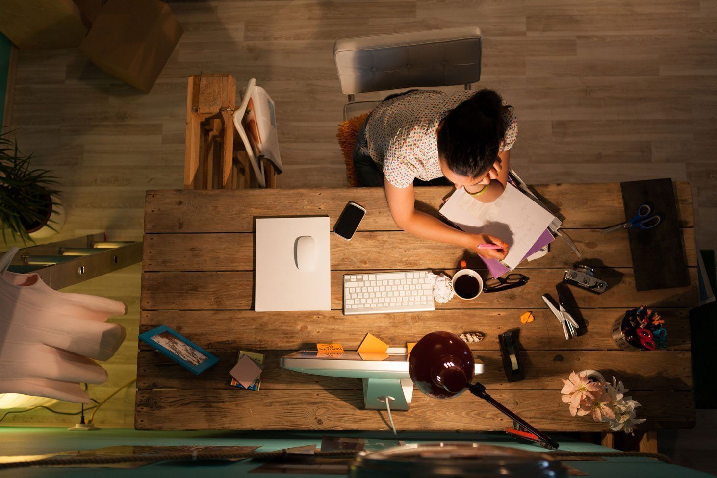 Overhead view of a person at a messy wooden desk, writing in a notebook; warm light, with phone and keyboard visible.