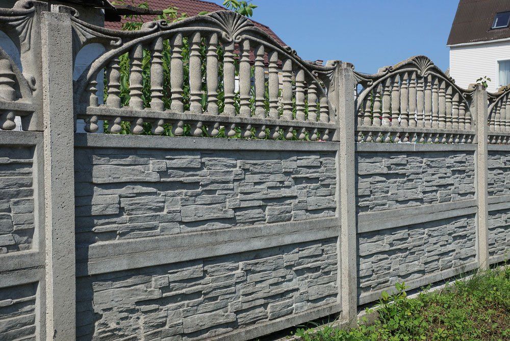 Gray concrete fence with decorative balusters and stacked-stone panels along a grassy edge under blue sky