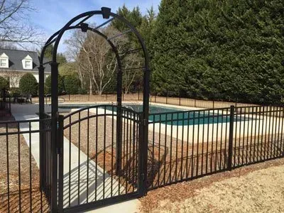 Black metal gate opening to an outdoor swimming pool surrounded by a fence and trees