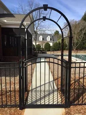 Black metal garden gate with arched trellis in front of a walkway and pool area