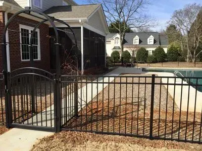 Black metal gate beside a brick house, opening to an empty backyard with a pool and trees beyond.