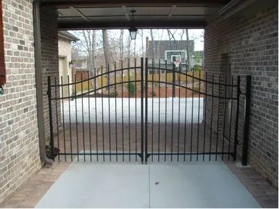 Black metal gate under a covered brick carport, blocking a driveway with a basketball hoop beyond.