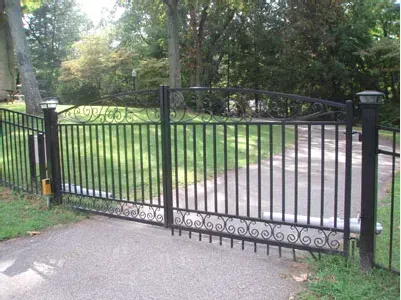 Black metal driveway gate across a paved entrance beside a grassy yard and trees
