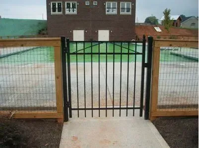 Black metal gate between wooden fences, opening to a paved courtyard with a brick building in the background