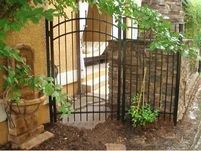 Arched black metal gate in a stone garden wall, opening onto a narrow stone path with plants nearby.