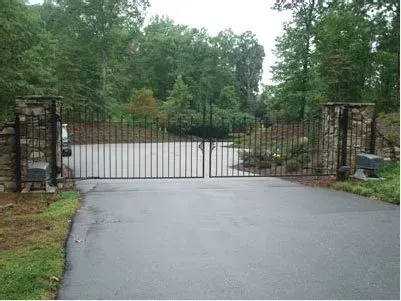 Closed black metal gate across a driveway between stone pillars and trees.