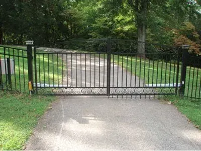 Black metal gate blocking a paved driveway, with trees and grass on both sides.