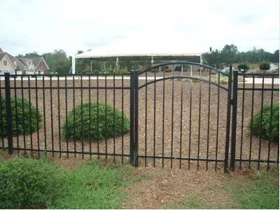 Black metal gate in front of a gravel lot with shrubs and houses in the background