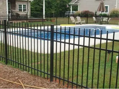 Backyard swimming pool enclosed by a black metal fence, with patio chairs and a house in the background
