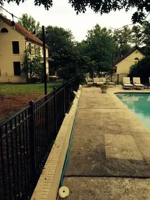 Poolside walkway beside a black fence, with lounge chairs, trees, and buildings under cloudy skies
