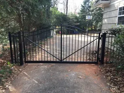 Black metal driveway gate at the end of a paved path, framed by trees and a house.