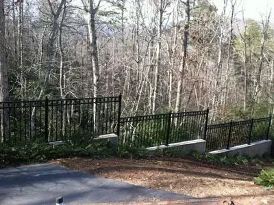 Black metal fence along a wooded hillside beside a paved road and concrete retaining wall