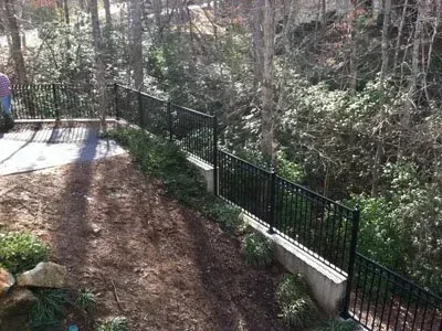 Wooded path beside a black metal fence and concrete retaining wall, with sunlight filtering through bare trees.