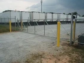 Closed chain-link gate at an industrial yard with white portable buildings and yellow bollards.