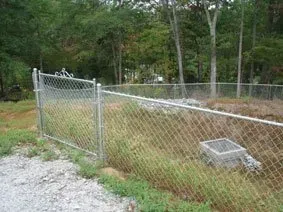 Chain-link fence around a grassy area with a concrete utility cover and trees in the background