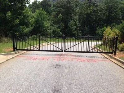 Black metal gate blocking a paved road, with dense green trees behind it.