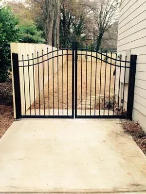 Black metal gate blocking a driveway, with a paved path and trees beyond