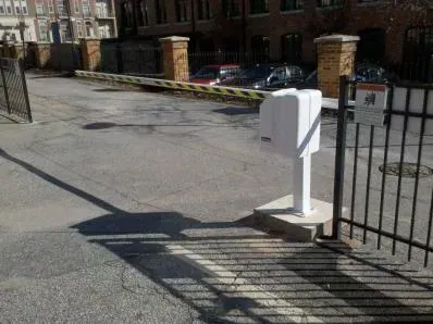 Driveway entrance with a white gate arm, black fence, and brick posts beside apartment buildings.