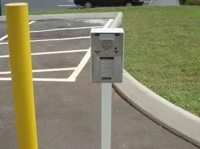 Parking lot access keypad on white post beside yellow bollard and curb.