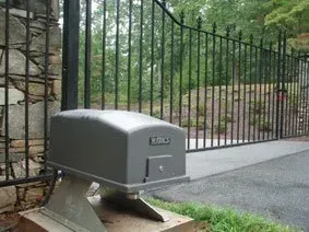 Gray automatic gate opener beside a black metal driveway gate and stone wall outdoors