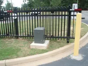 Black metal fence beside a curb with a gray utility box and yellow bollard in a grassy area