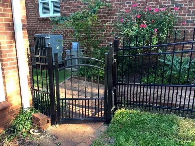 Black metal gate opening onto a brick garden path beside a brick house and green lawn.