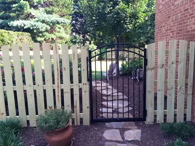 Black garden gate in a pale wooden fence, with stone path leading into a leafy yard.