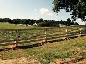 Wooden fence along a grassy field under a sunny sky, with trees and a farmhouse in the distance