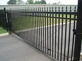 Black metal swing gate across a driveway and sidewalk, with vertical bars and pointed finials.