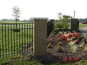 Black metal fence beside brick pillars and a landscaped flower bed in a grassy park.