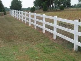 White wooden fence running along a grassy field beside a dirt road, with trees in the distance