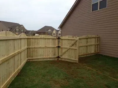 Wooden backyard fence with a gate beside a house, enclosing a grassy corner yard.