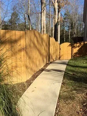 Curving concrete path beside a tall wooden fence in a wooded backyard on a sunny day