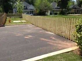 Driveway beside a long wooden fence, with a lawn and houses in the background.