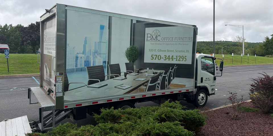 White box truck with city skyline graphic parked in a lot beside bushes and a grassy field
