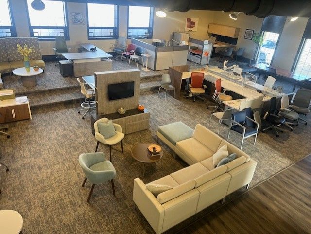 Office space with rows of desks, chairs, and plants under white ceiling lights.