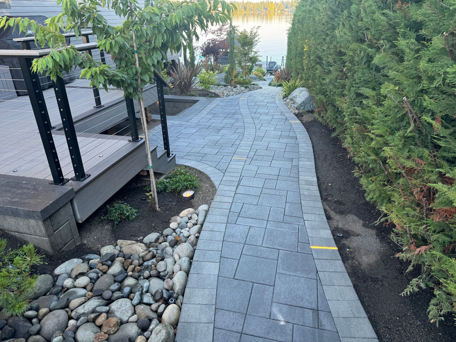 A gray home with a paved pathway leading from a dock on a lake, surrounded by green grass and trees.