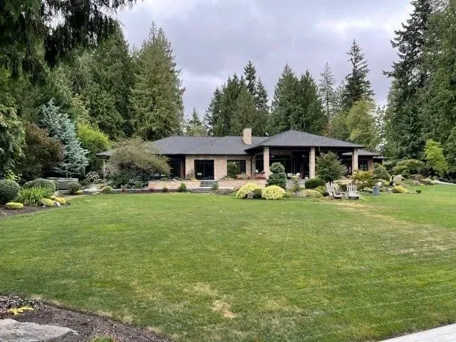 House with large lawn, surrounded by trees. Overcast sky.