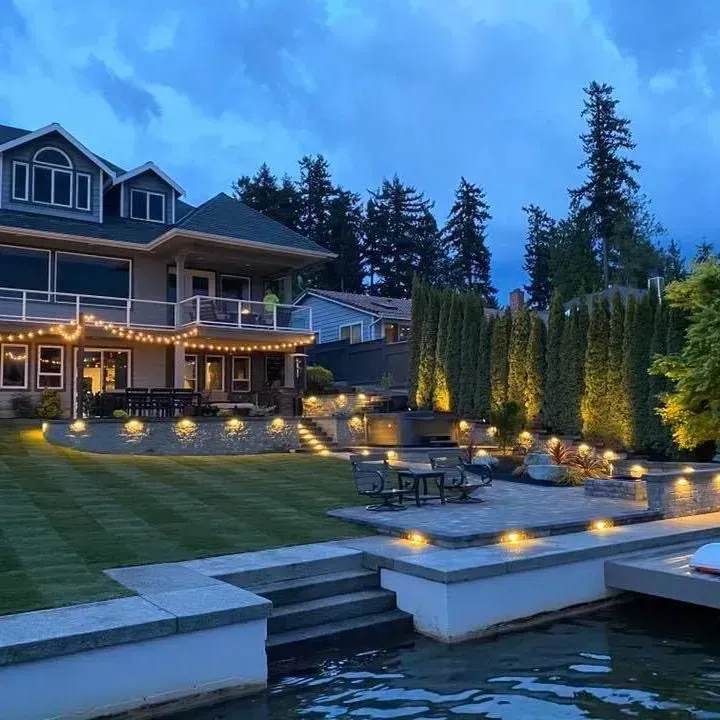 Large lakeside home with tiered patio and landscape lighting at dusk.