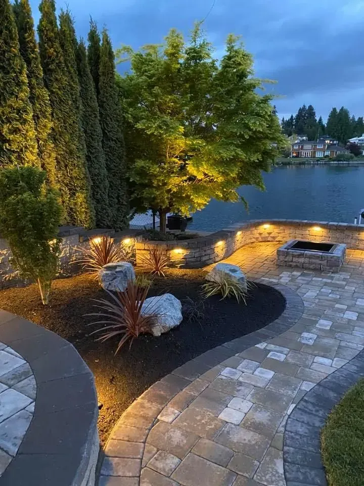 Landscaped patio with stone wall, lake view, and illuminated trees at dusk.