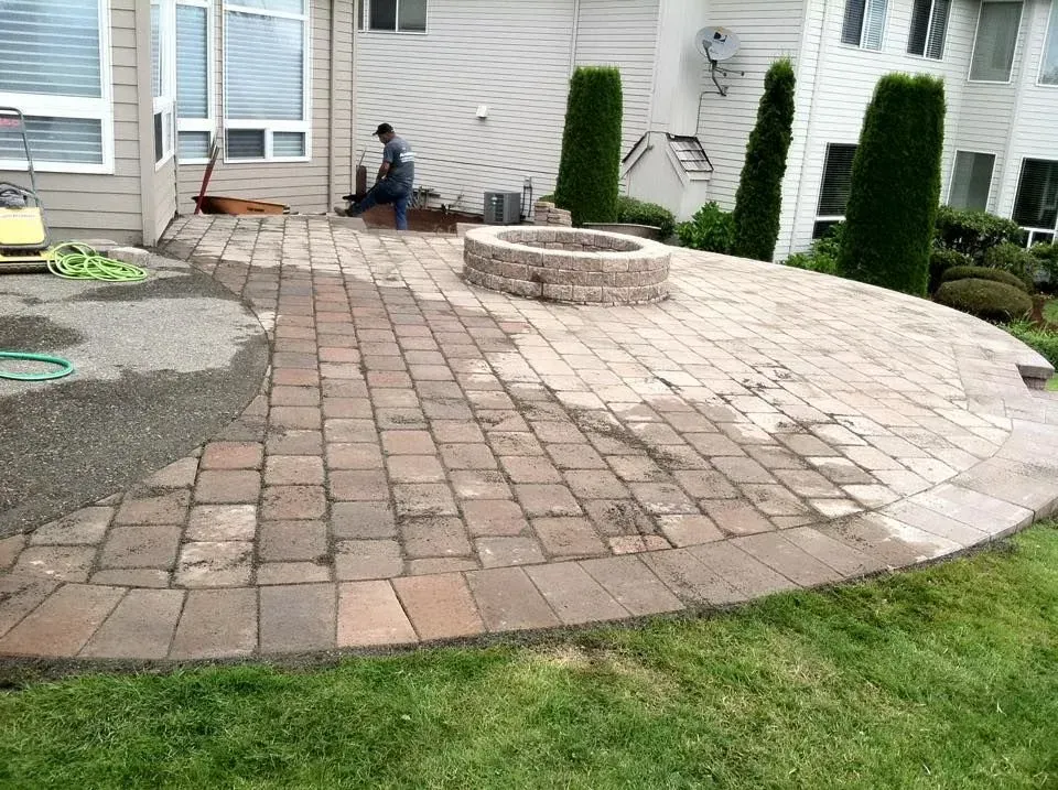 Brick patio with a fire pit, bordered by grass. A person works nearby.