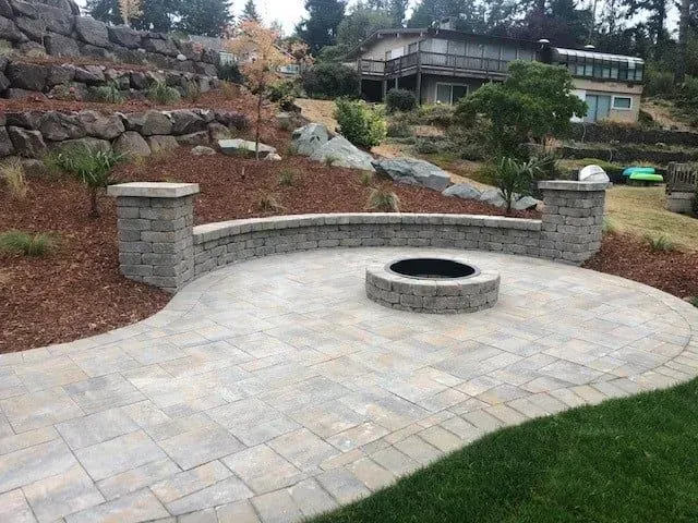 Patio with fire pit, curved retaining wall, and stone columns, overlooking terraced yard and house.