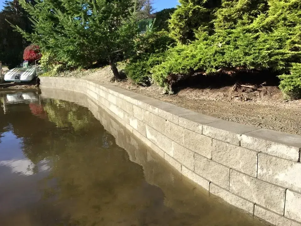 A concrete retaining wall lines a pond, with greenery in the background.