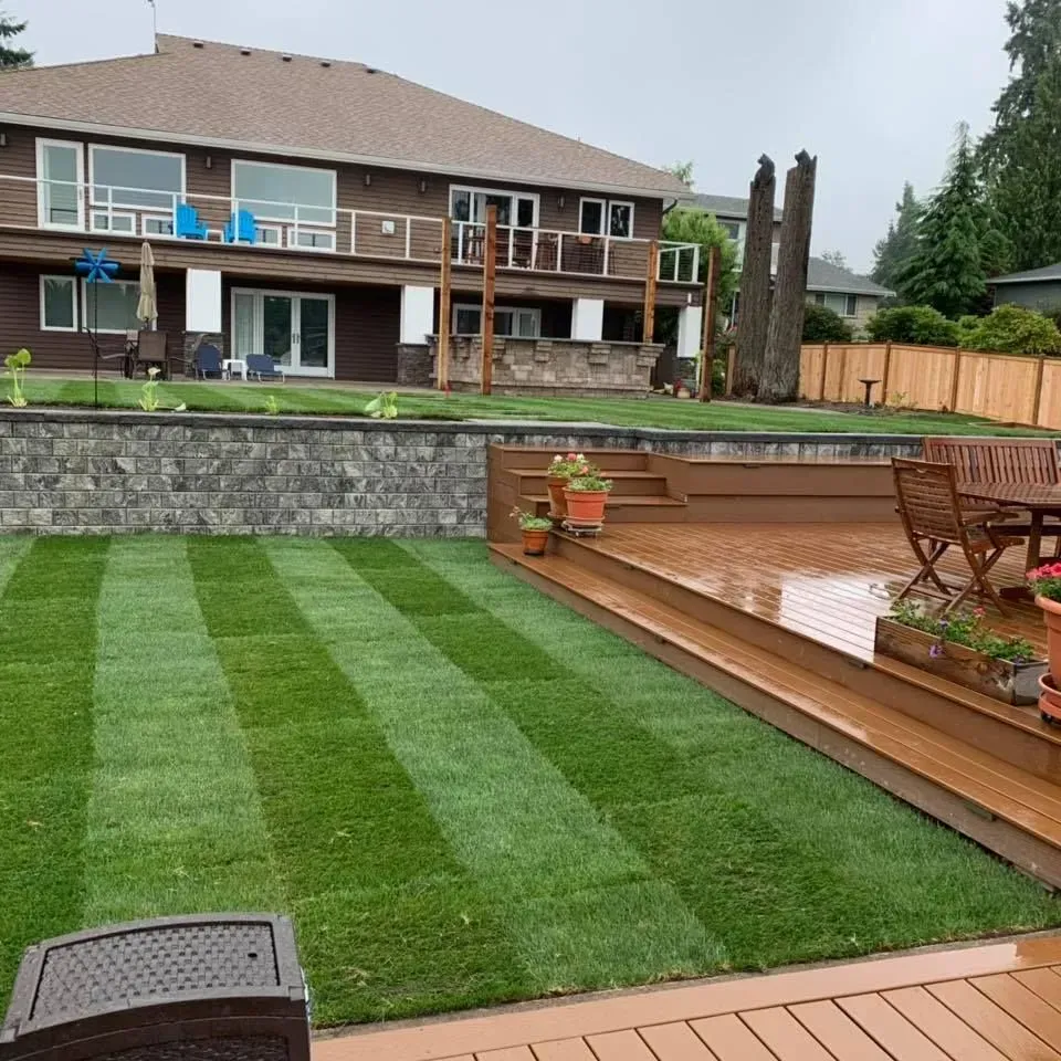 Green lawn striped with mowing in a backyard with a house, deck, and stone wall.