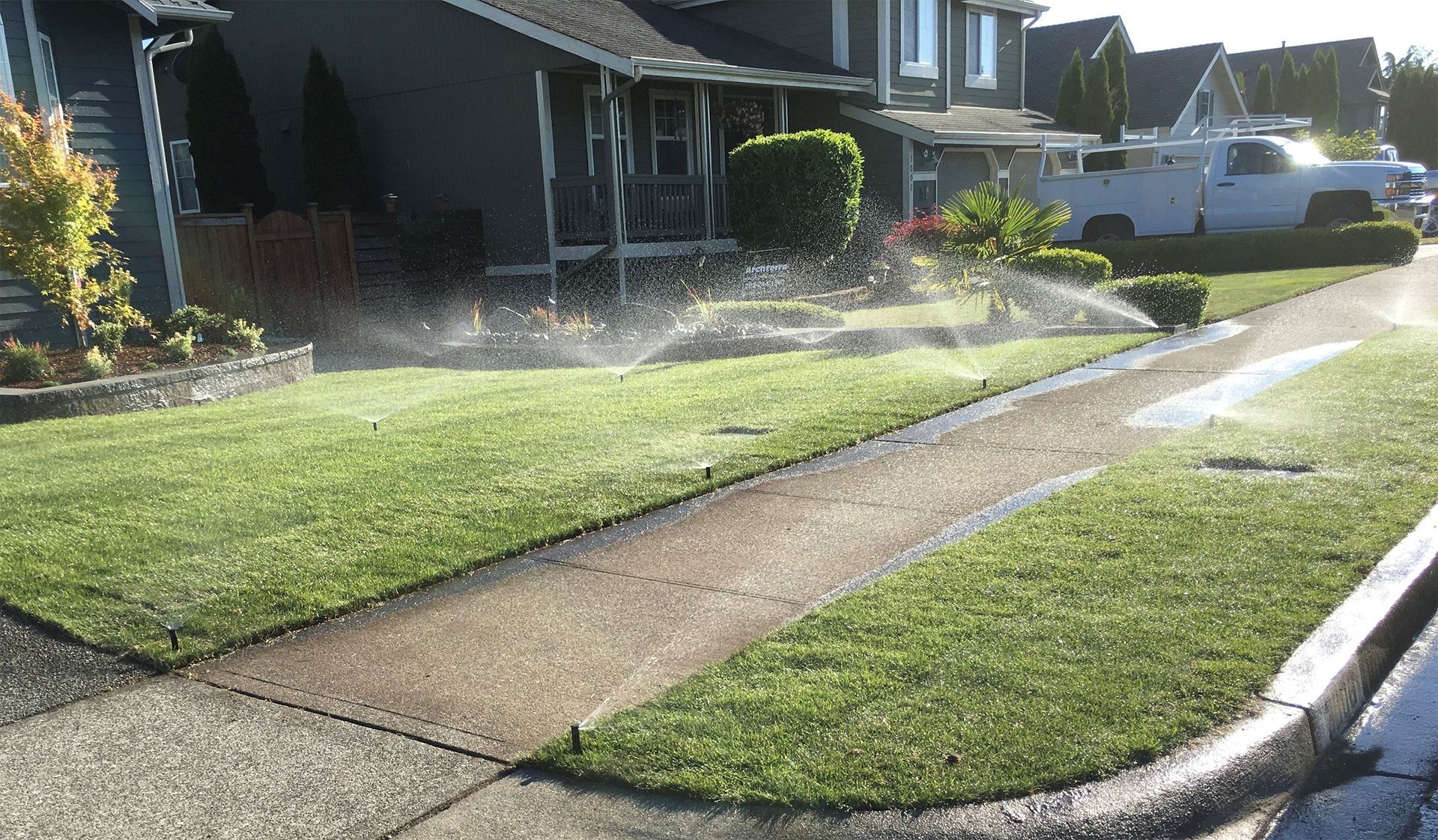 Sprinklers watering green lawn in front of houses; sidewalk in foreground; sunny day.