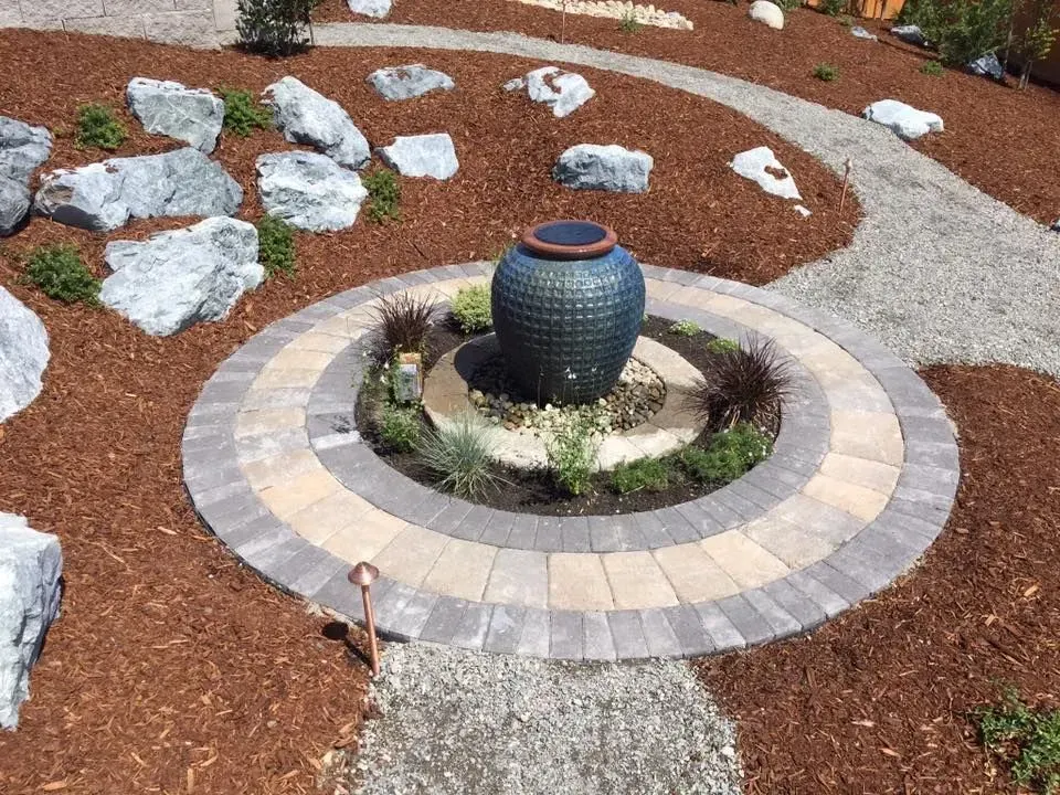 Fountain with blue glazed pot in circular brick design, surrounded by mulch and rocks, garden path.