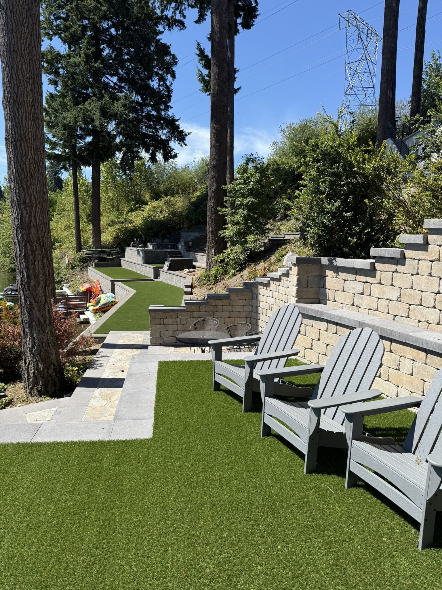 Lawn chairs on artificial turf next to a stone wall and pathway with trees and a sunny sky.
