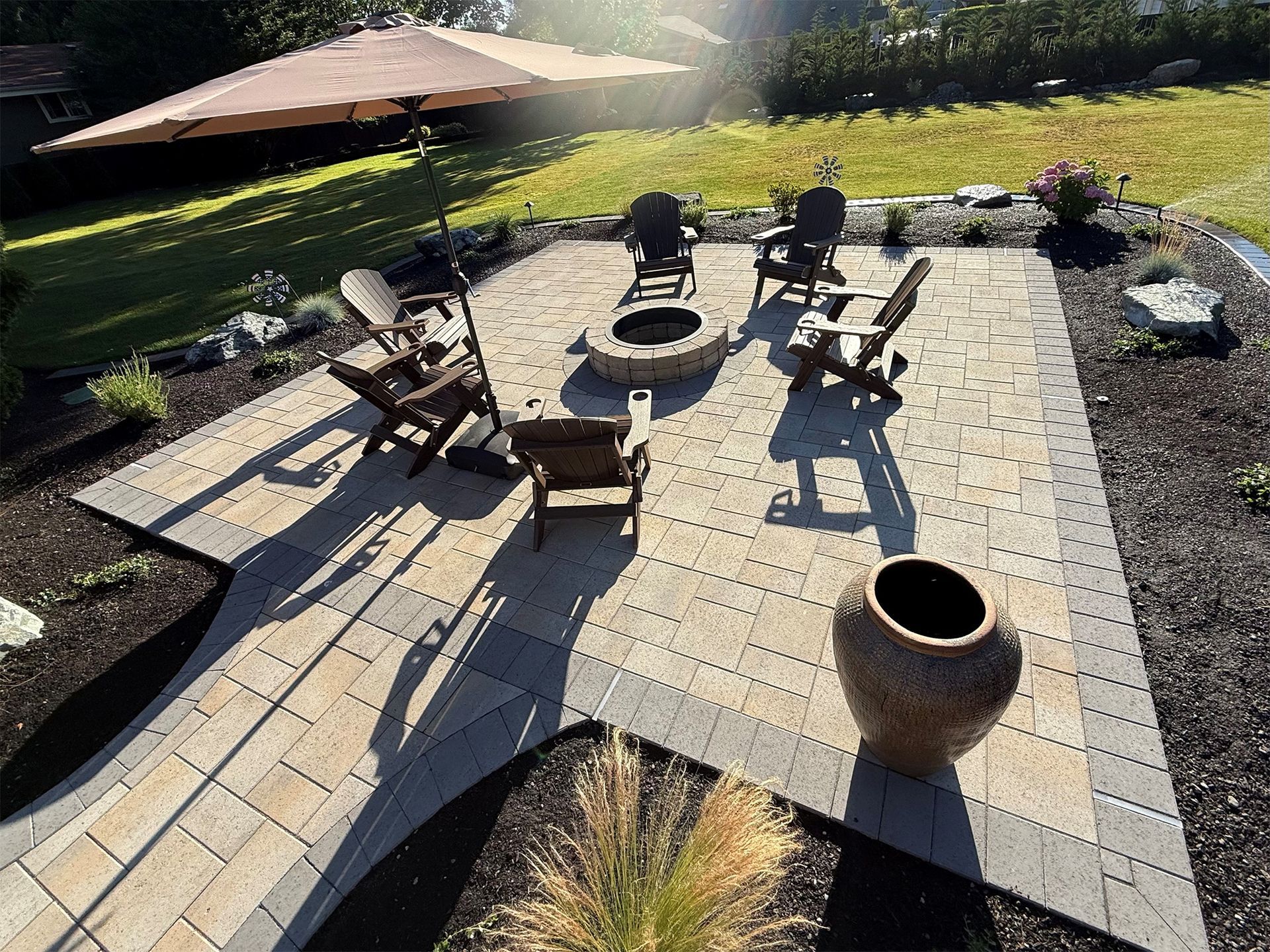 Patio with fire pit, chairs, umbrella, and decorative urn. Grass and landscaping surround the brickwork.