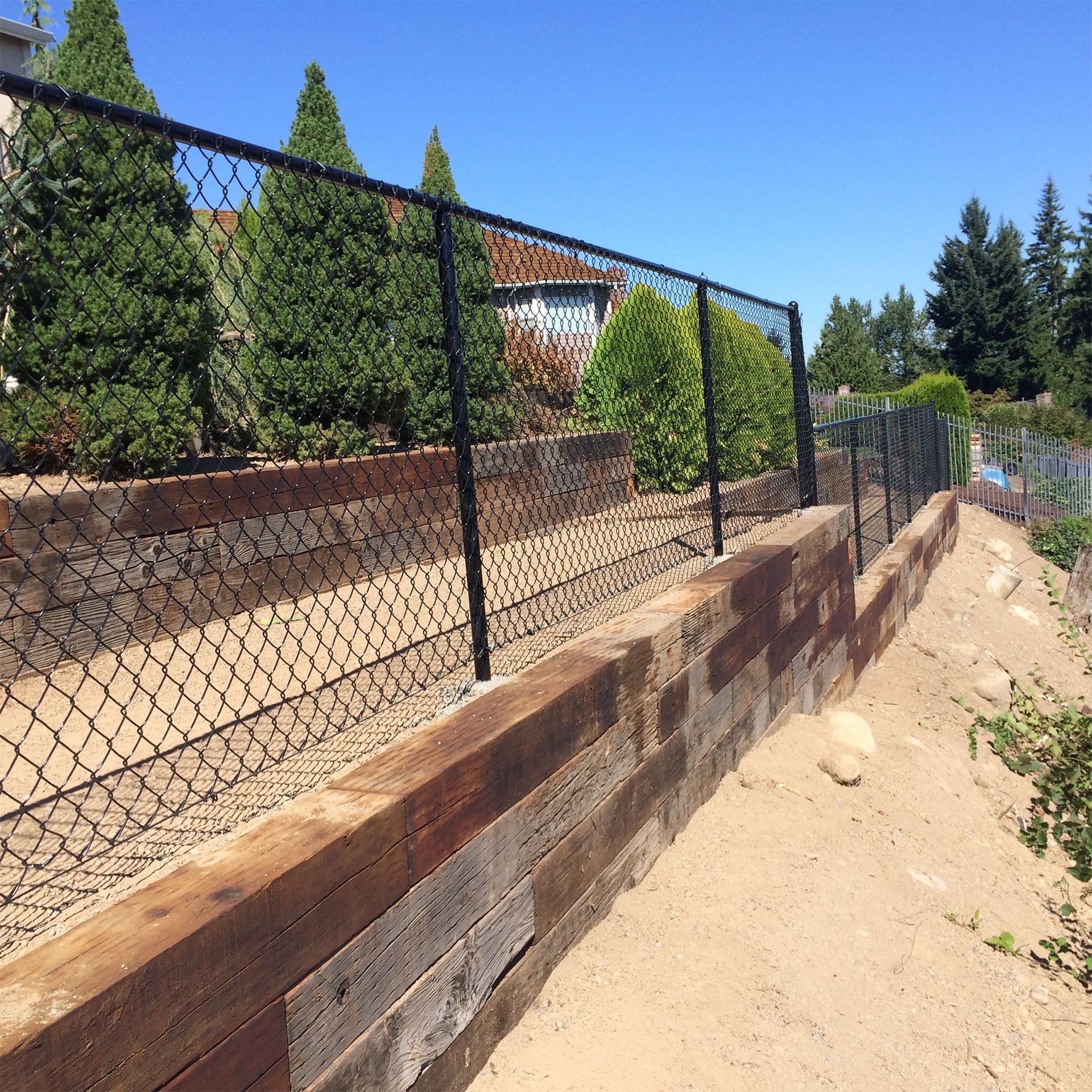 Black chain-link fence atop a weathered wood retaining wall in a yard with trees and sandy ground.