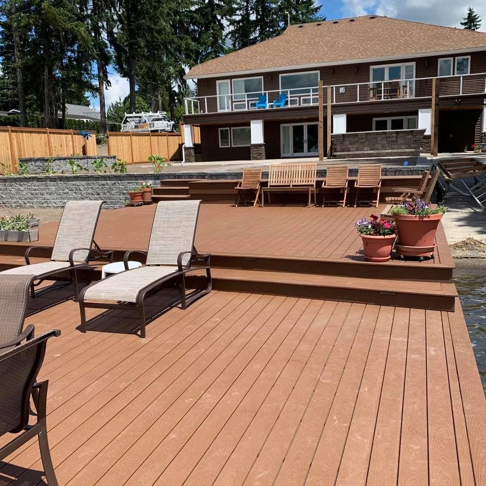 Lakeside deck with lounge chairs, overlooking water. Brown house with balcony. Sunny day.